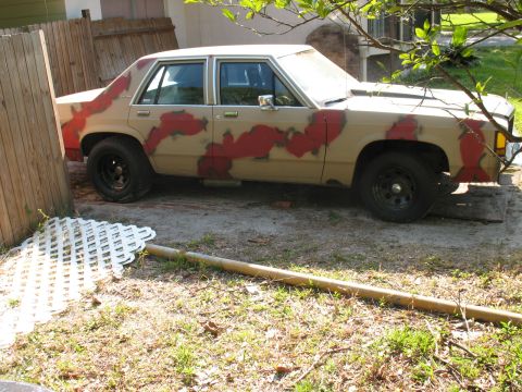 1985 Ford LTD Crown Victoria - A tan sedan with a crude red camouflage paint job sits in a yard next to a wooden fence.