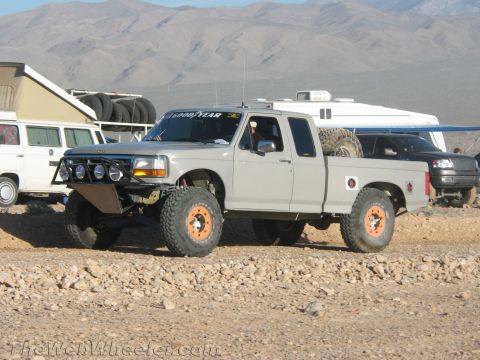 1990 Ford Bronco - silver Ford extended-cab pickup with a prerunner front bumper, auxiliary lights and orange beadlock wheels sits at a dusty desert staging a…