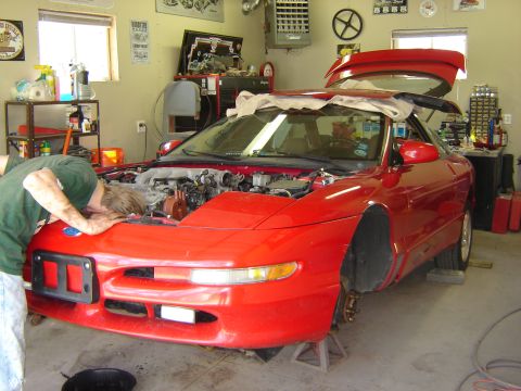 1993 Ford Probe - A person works on the engine of a red 1993 Ford Probe, which is raised on jack stands in a garage.