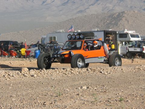 1990 Ford Bronco - orange and black multi-seat off-road buggy with a roof light bar drives across rocky desert ground past RVs and a US flag.