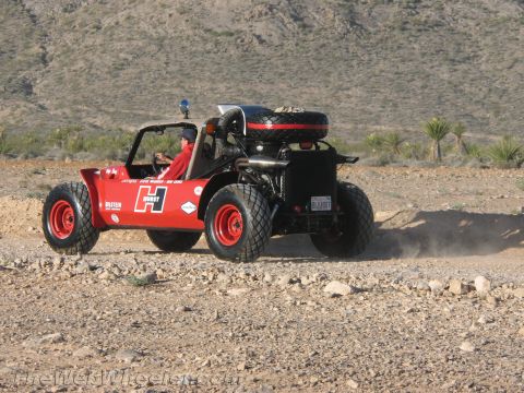 1990 Ford Bronco - red and black single-seat off-road buggy marked with a Hurst H logo and a roof-mounted spare tire kicks up dust on a desert trail.