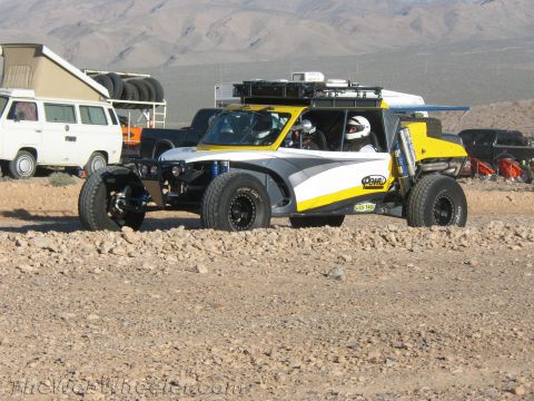 1990 Ford Bronco - white and yellow long-travel off-road buggy carrying two helmeted occupants sits on rocky desert ground among parked vehicles.