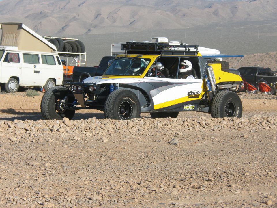 White and yellow long-travel off-road buggy with two helmeted occupants on rocky desert ground