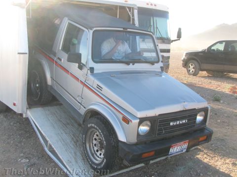 1990 Ford Bronco - silver Suzuki Samurai with red side stripes is driven up the ramp into an enclosed trailer at a dusty desert site.