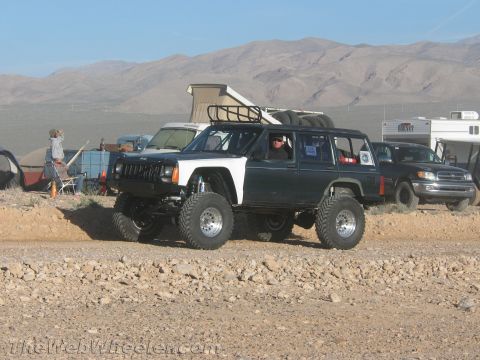1990 Ford Bronco - green and white lifted Jeep Cherokee with a roof basket sits on oversized tires at a dusty desert campsite with tents and RVs behind.