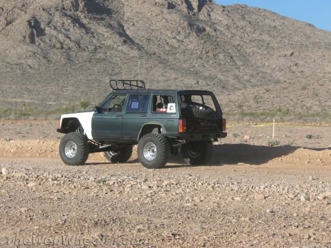 1990 Ford Bronco - Rear three-quarter view of a green and white lifted Jeep Cherokee with a roof basket and rear-mounted spare on a rocky desert trail.
