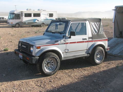 1990 Ford Bronco - silver Suzuki Samurai 4x4 with a black soft top and red side stripes sits parked beside motorhomes on dusty desert ground.