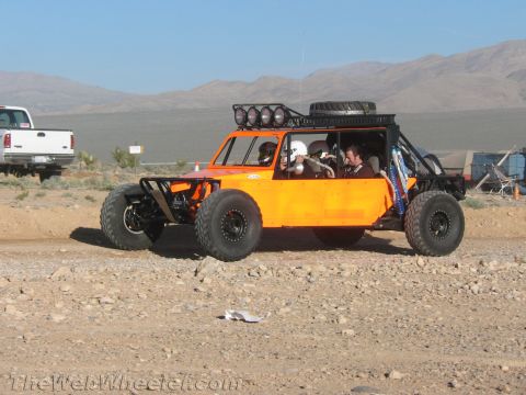 1990 Ford Bronco - orange long-wheelbase off-road buggy with a roof light bar and spare tire carries occupants along a dirt road in open desert.