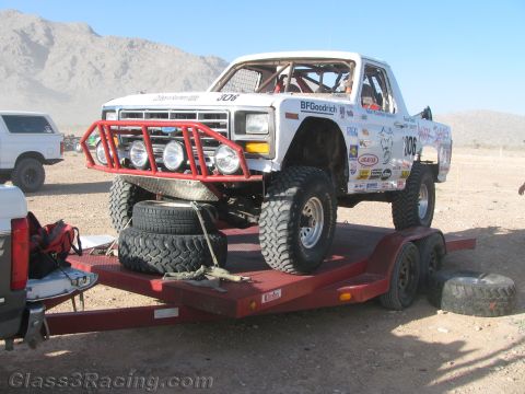 1990 Ford Bronco - white vintage Ford race pickup numbered 306 with a red tubular brush guard and BFGoodrich decals sits chained on a flatbed trailer with spa…