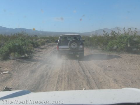 1990 Ford Bronco - Driver's-eye view through a dirty windshield of a dark SUV with a rear-mounted spare kicking up dust on a desert two-track road.