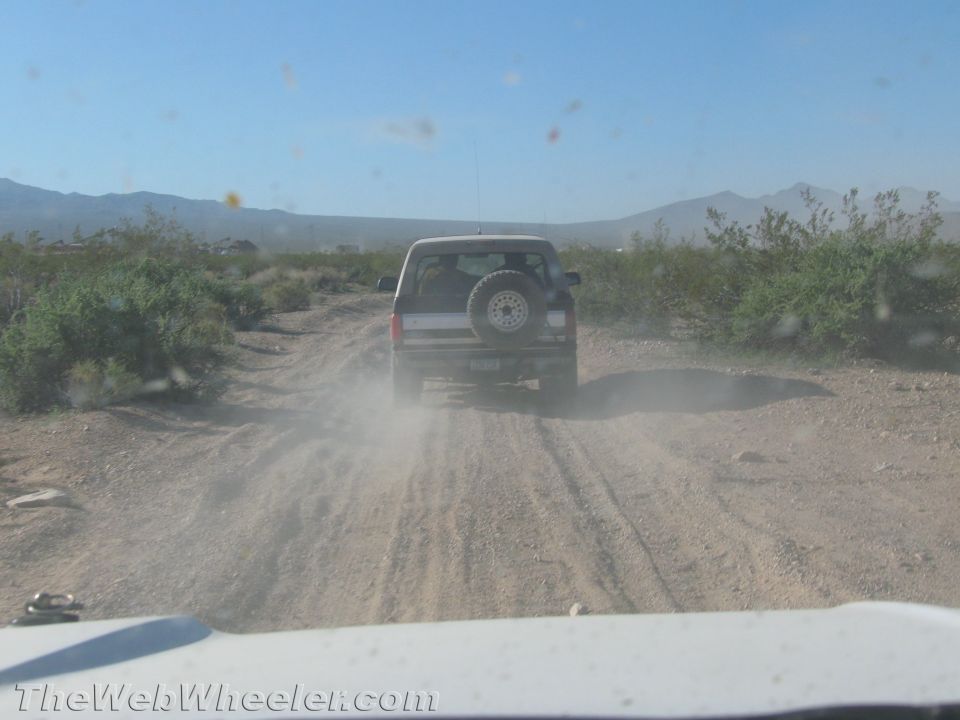 View through windshield of a dark SUV with rear spare tire driving ahead on a dusty desert road