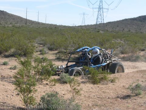 1990 Ford Bronco - blue tube-frame single-seat dune buggy with a helmeted driver sits in open desert scrub under power lines.