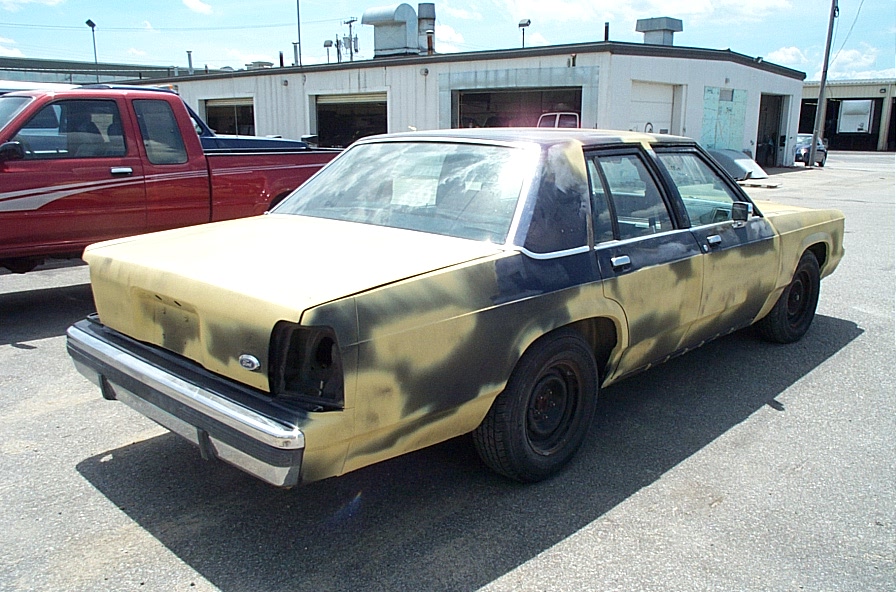 Yellow-primer four-door sedan on steel wheels in a shop lot, rear 3q view