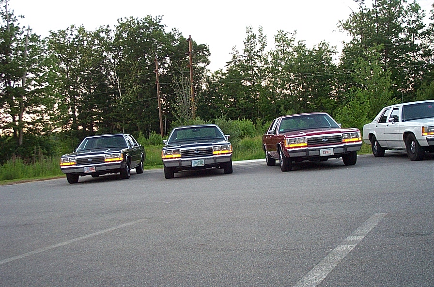 Row of four boxy 1980s Ford and Mercury full-size sedans parked in a lot at dusk