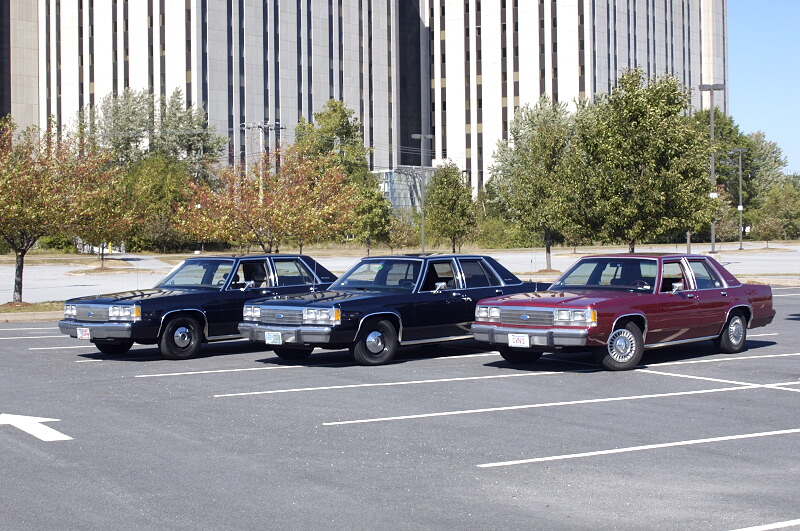 Three boxy Ford full-size sedans, two dark and one maroon, parked in a lot by an office tower