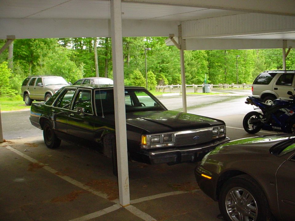 Black Ford sedan parked under carport, front 3/4 view with SUVs and a Yamaha motorcycle nearby.