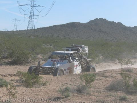 1990 Ford Bronco - white tube-frame off-road race buggy speeds along a dusty desert trail beneath transmission towers.