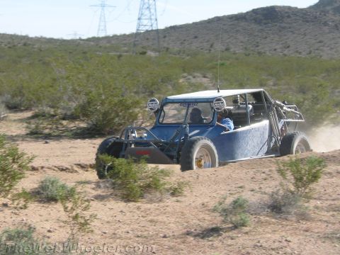 1990 Ford Bronco - blue-and-white four-seat off-road buggy with roof lights races across sandy desert brush.