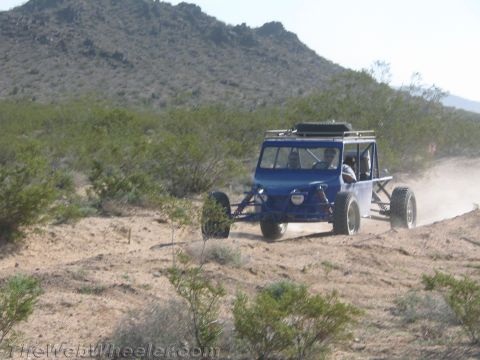 1990 Ford Bronco - blue tube-frame off-road buggy with a roof rack drives along a sandy desert trail near a rocky hillside.