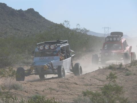 1990 Ford Bronco - white buggy lettered Dirt Mob leads a red off-road vehicle along a dusty desert hill trail.