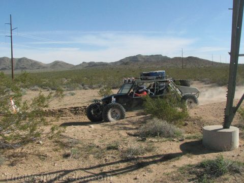 1990 Ford Bronco - long-wheelbase tube-frame off-road buggy loaded with gear crosses desert scrub past a utility pole.