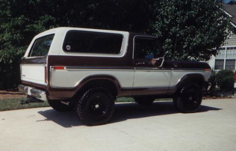 1978 Ford Bronco - A two-tone brown and white 1978 Ford Bronco is shown from the side, parked on a concrete surface with trees and a house in the background.