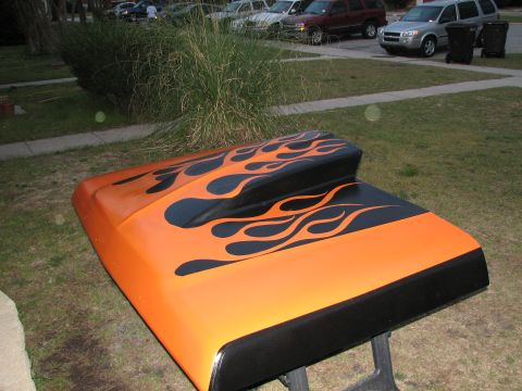 1979 Ford Bronco - An orange hood with a raised cowl scoop, finished in black tribal flames and a solid black front band, resting on sawhorses on a lawn. Park…