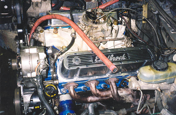 Blue V8 engine with Edelbrock valve cover, carburetor, headers and red hose in an engine bay
