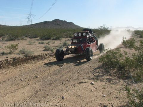 1990 Ford Bronco - red tube-frame off-road buggy with roof light bar throws dust along a sandy desert two-track trail.