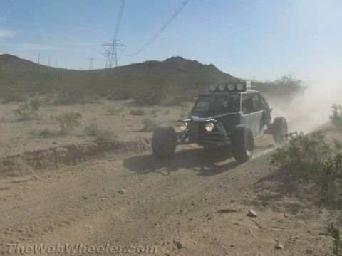 1990 Ford Bronco - dark tube-frame off-road buggy with headlights and a roof light bar speeds along a dusty desert trail.