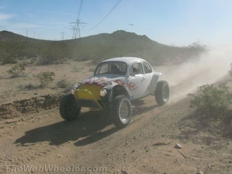 1990 Ford Bronco - white Baja-style VW Beetle with yellow-and-red flame paint races down a dusty desert trail.