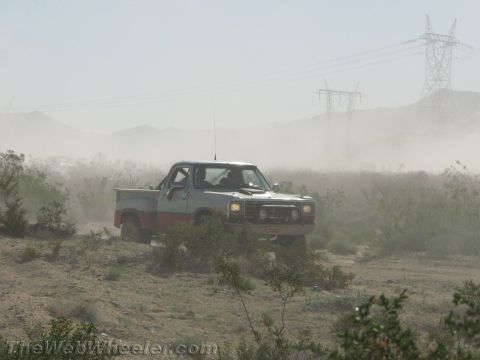 1990 Ford Bronco - older silver-and-red Dodge pickup with headlights on plows through dust in open desert scrub.