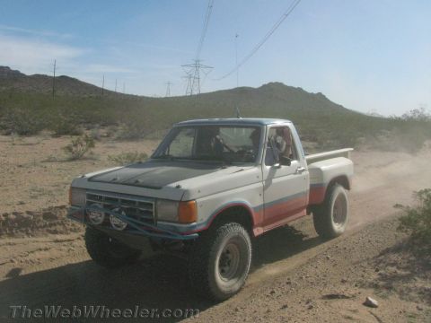 1990 Ford Bronco - older white-and-multicolor Ford pickup with an off-road light bumper drives a sandy desert trail.