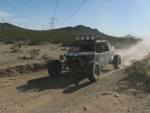 1990 Ford Bronco - silver tube-frame off-road buggy with a roof light bar throws dust on a sandy desert trail.