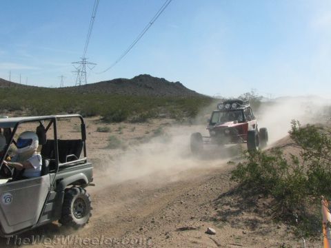 1990 Ford Bronco - From a gray side-by-side UTV, a red off-road buggy is seen racing up a dusty desert hill trail.