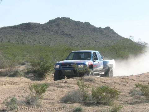 1990 Ford Bronco - lifted multicolor Toyota pickup truck races across desert brush below a rocky hillside.
