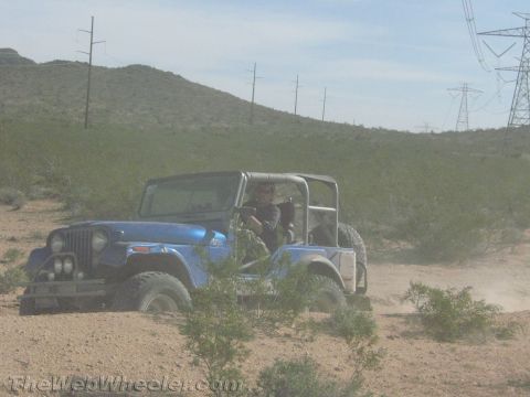 1990 Ford Bronco - blue open-top Jeep CJ on large off-road tires drives through dusty desert brush with a driver aboard.