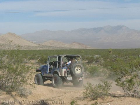 1990 Ford Bronco - blue Jeep CJ with a rear-mounted spare tire crosses a sandy desert trail with mountains behind.