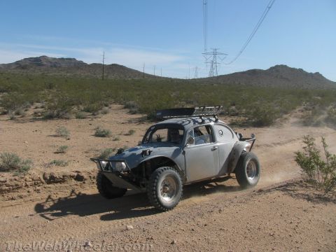 1990 Ford Bronco - bare-metal Baja-style VW Beetle with a roof basket drives a sandy desert trail under power lines.