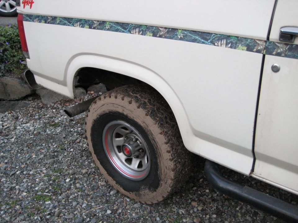 White SUV with camouflage stripe and 4x4 decal on rear quarter panel. Muddy tire and exhaust pipe visible