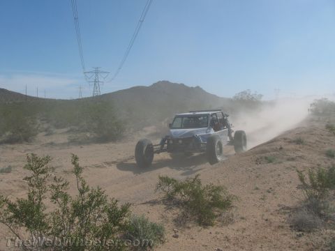 1990 Ford Bronco - white tube-frame off-road buggy climbs a dusty desert berm beneath transmission towers.