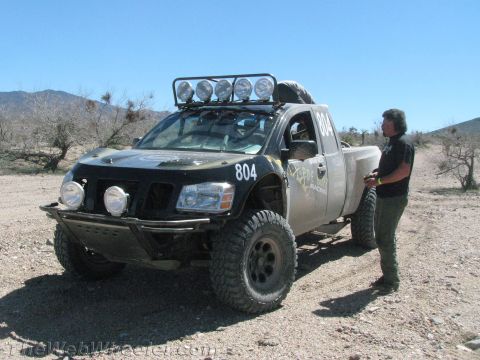 1990 Ford Bronco - person stands beside a black prerunner pickup truck numbered 804 with a roof light bar in the desert.