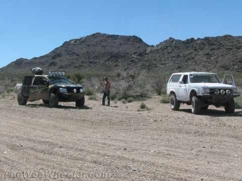 1990 Ford Bronco - black race truck and a white off-road SUV sit on a dirt road in the open desert with a rocky hill behind them.