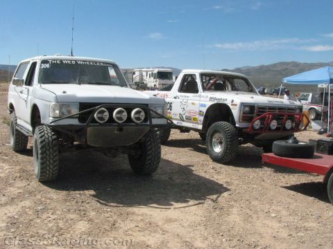 1990 Ford Bronco - white off-road SUV with quad headlights and a white sponsored race truck numbered 306 are staged at a dusty desert pit.