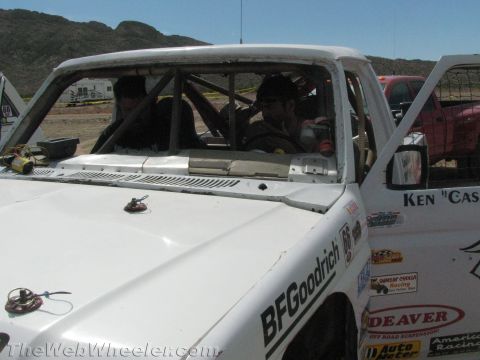 1990 Ford Bronco - driver sits inside the roll-caged cab of a white BFGoodrich-sponsored off-road race truck at a desert pit.