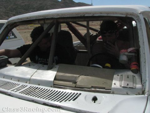 1990 Ford Bronco - Two people sit inside the roll-caged cab of a white off-road race truck, seen across the vented hood in the desert.