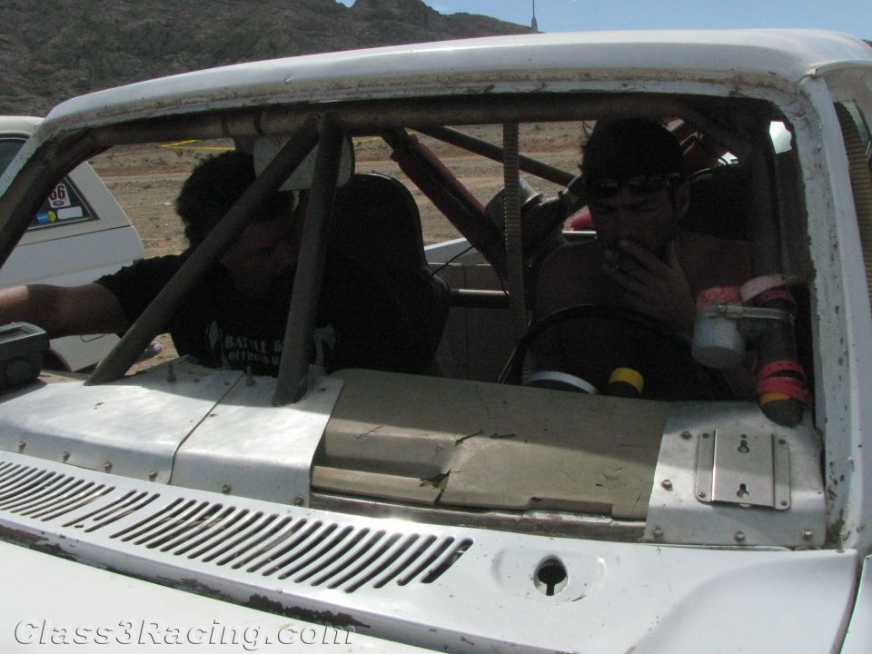 Two people inside roll cage of white race truck viewed over the hood