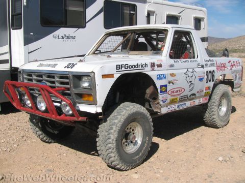 1990 Ford Bronco - white Ford off-road race truck numbered 306 with a red tube bumper and sponsor decals sits beside a motorhome in the desert.