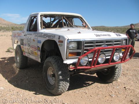 1990 Ford Bronco - white Ford off-road race truck numbered 306 with red tube bumper and sponsor decals sits on desert ground near a person.