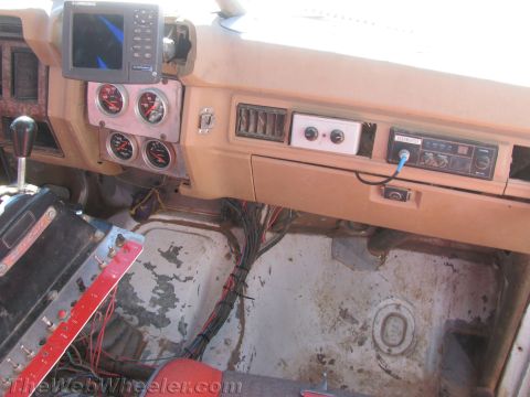 1990 Ford Bronco - Interior of a race vehicle showing a tan dashboard with auxiliary gauges, a stereo, a GPS unit and exposed wiring over a bare floor.